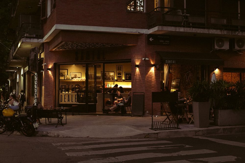 Fachada nocturna de SĀNTAL Colegiales en Conde 1200 esquina Virrey Loreto, con mesas en la vereda